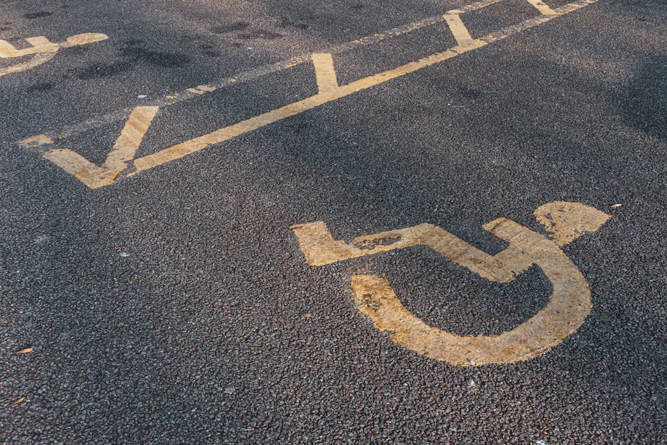 Close-up image of a designated accessible parking space sign painted on a dark asphalt surface, with faded white paint showing signs of wear and weathering. The parking space lines are visible in the