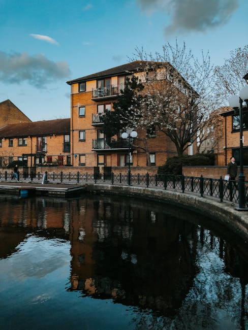 Photograph of a multi-storey brick residential building adjacent to a waterway, with a paved walkway and black metal railing along the edge. Several residents are visible walking along the pavement, d